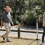 A man in a hat strolls on green grass during a 1 Day Home Away Tour, with outdoor kit and lush trees visible in the background.