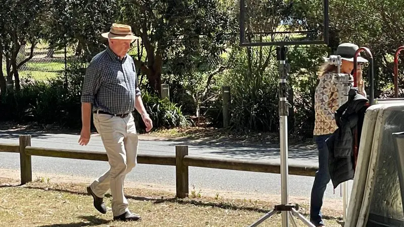 A man in a hat strolls on green grass during a 1 Day Home Away Tour, with outdoor kit and lush trees visible in the background.