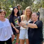 A joyful group poses for a selfie taken by two women during a 1 Day Home Away Tour, surrounded by lush trees and glowing sunlight.