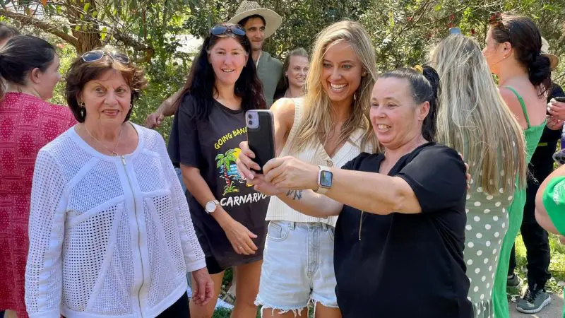 A joyful group poses for a selfie taken by two women during a 1 Day Home Away Tour, surrounded by lush trees and glowing sunlight.