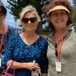 Three happy women on a Celebtime Tours 1 Day Home Away Tour, posing outside amid lush greenery and a scenic building backdrop.