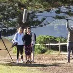 A professional film crew captures a 1 Day Home Away Tour with two participants outdoors, surrounded by lush trees and a wooden fence.