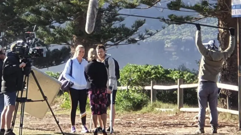 A professional film crew captures a 1 Day Home Away Tour with two participants outdoors, surrounded by lush trees and a wooden fence.