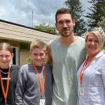 Group of four wearing orange lanyards stands outdoors on a 1 Day Home Away Tour, smiling by a building surrounded by green trees.