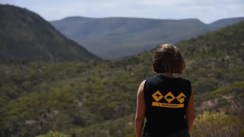 Traveller in a black "I Survived" shirt overlooks lush green hills during the 10-Day Adelaide to Perth Adventure Tour with Untamed Escapes.