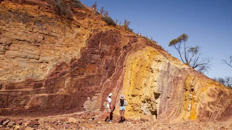 Explore the stunning West MacDonnell Ranges on a 1 Day Standley Chasm tour; two people stand by layered rocks beneath blue skies.