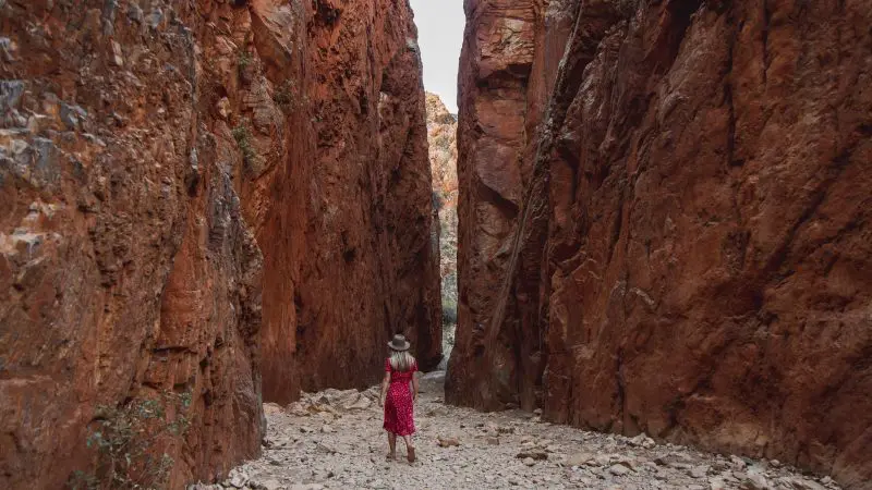 A woman in a vibrant red dress hikes through Standley Chasm’s striking, narrow red rock cliffs on a West MacDonnell Ranges day tour.