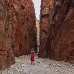 Experience the breathtaking 4 Day Uluru to West MacDonnell Tour as a woman in a red dress explores vibrant red canyon cliffs.