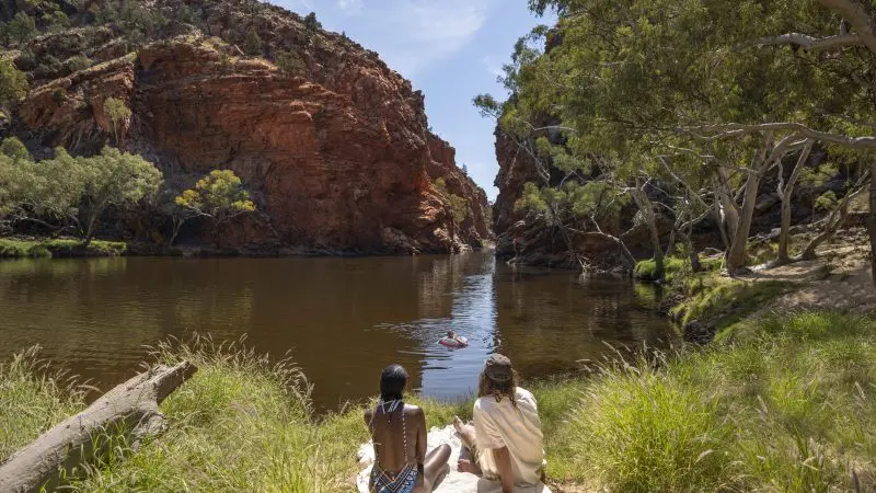 Experience a scenic 1 Day West MacDonnell Ranges tour: two people relax riverside beneath striking red cliffs near Standley Chasm.