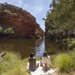 Two travellers relax beside a tranquil river, admiring vibrant red cliffs on a guided 4-Day Uluru to West MacDonnell tour, blue sky overhead.