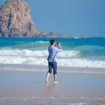 Barefoot visitor enjoys panoramic sea views at Phillip Island Penguin Parade as playful koalas wander nearby on the sandy beach.