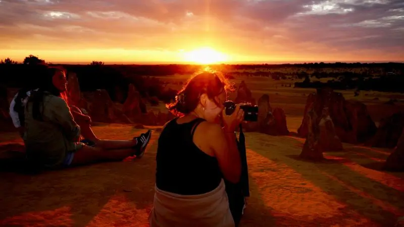 Two travellers enjoy a vibrant sunset atop rocky terrain on an Autopia 1 Day Pinnacles Sunset Stargazing Tour as one takes photos.