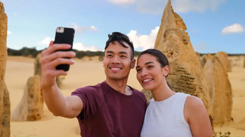 Happy couple takes a selfie at Pinnacles Desert during their Autopia 1 Day Sunset Stargazing Tour, capturing golden dunes at dusk.