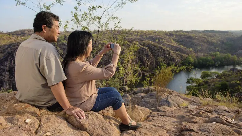 Couple relax on rocky terrain, documenting their 1 Day Katherine Gorge Cruise amid stunning canyon views and a winding river backdrop.
