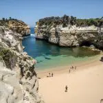 Stunning sandy beach with vibrant turquoise water, dramatic cliffs, and clear blue sky on Great Ocean Road Sunset Tour from Melbourne.
