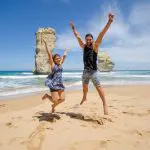 Two people jump joyfully on a sandy beach with waves and rocks, enjoying a 1 Day Great Ocean Road Rainforest Tour adventure.