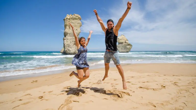Two people jump joyfully on a sandy beach with waves and rocks, enjoying a 1 Day Great Ocean Road Rainforest Tour adventure.
