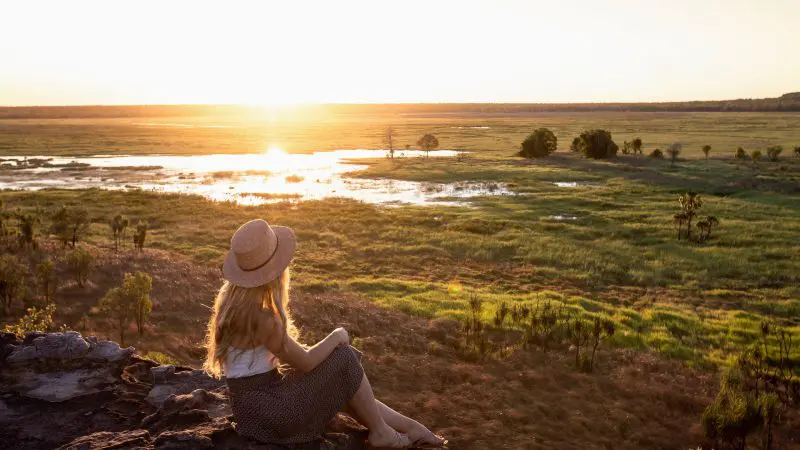 Woman wearing a hat relaxes on a rock at sunset, taking in the breathtaking Kakadu Outback landscape on a 2 Day Retreat Tour from Darwin.