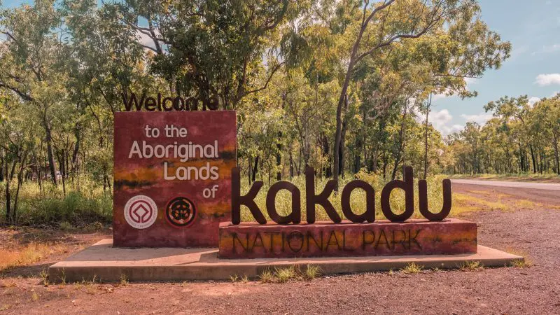 Entrance sign greets 2 Day Kakadu Outback Retreat Tour From Darwin guests to Aboriginal Lands, with lush trees and road in background.