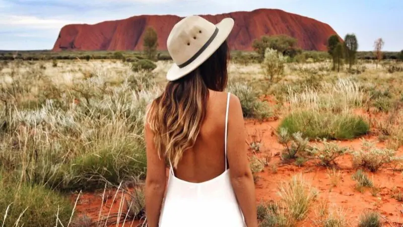 Woman in white dress and hat standing before iconic Uluru during a 3-Day Red Centre Kings Canyon Safari from Alice Springs, Australia.