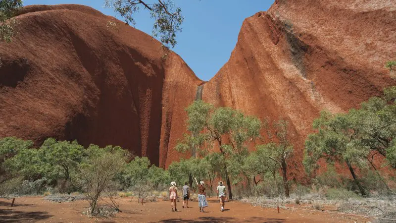 Travellers on a 3 Day Uluru Kings Canyon Safari from Alice Springs approach iconic Uluru’s base, beneath the clear blue Australian sky.