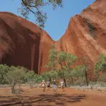 Tour group approaches Uluru on a 4-Day Uluru Kata Tjuta Kings Canyon adventure, blue sky above stunning Australian outback landmarks.