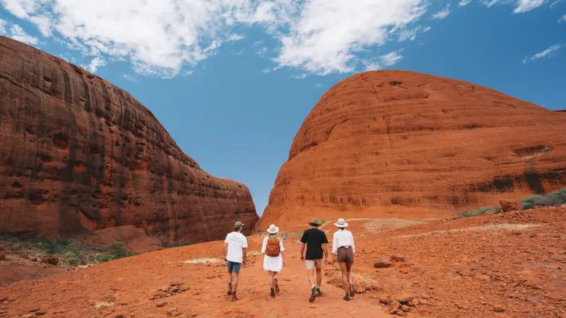 Four travellers hiking a vibrant red desert trail on a 2 Day Red Centre Rock Tour from Alice Springs, beneath stunning sandstone formations.