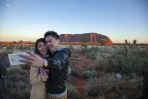 A smiling couple snaps a sunset selfie at Uluru on their 2 Day Uluru Kata Tjuta Rock Tour from Ayers Rock Yulara, iconic outback view.