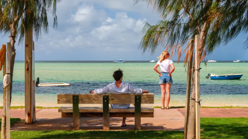Two people on a Perth to Exmouth coastal tour: one seated on a bench, another standing by the sea, admiring boats and ocean views.