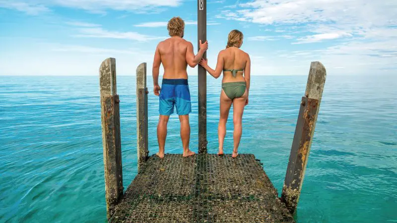Two travellers in vibrant swimwear stand on a rustic dock, envisioning an epic 6 Day Perth to Exmouth Coral Coaster adventure tour.