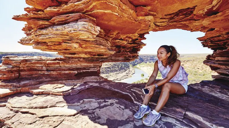 A woman relaxes on rocky terrain beneath a scenic stone arch, gazing at a river during her 6 Day Perth to Exmouth Coral Coaster tour.