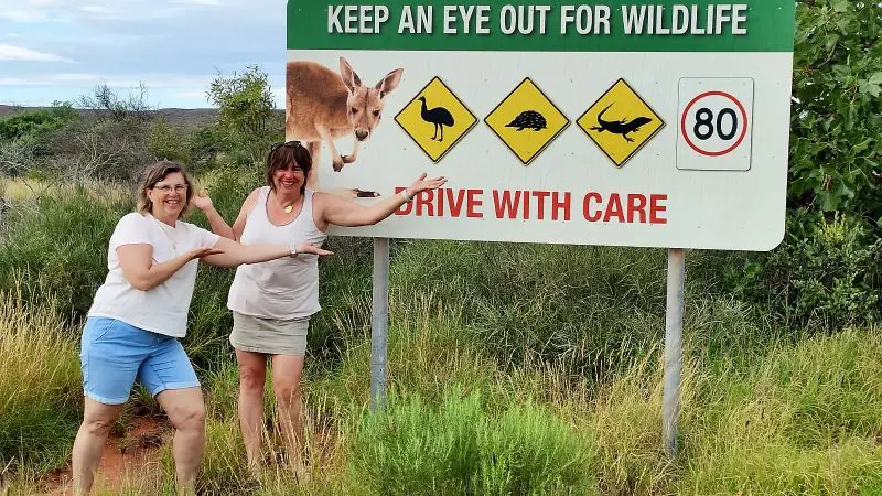 Two smiling women point at a wildlife warning sign on a scenic 4 Day Perth to Monkey Mia Coastal Loop Return Tour, perfect for travellers.