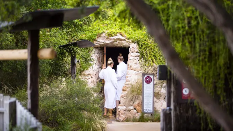 Two guests in white dressing gowns enter a luxurious stone sauna at Peninsula Hot Springs after arriving via the 1 Day Spa Entry Shuttle Bus.