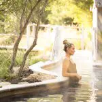 A woman in a stylish black swimming costume unwinds in a natural hot spring after arriving via the Peninsula Hot Springs Spa Entry Shuttle Bus.