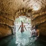 Experience rejuvenation as a woman strolls through a cave’s stone arch and man unwinds nearby; Peninsula Hot Springs shuttle bus outside.