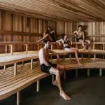 Four guests in swimwear unwind on benches inside a contemporary sauna after enjoying a Peninsula Hot Springs Spa Entry Shuttle Bus experience.