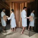 Two guests in white robes enter a wooden sauna at Peninsula Hot Springs, holding 1 Day Spa Entry shuttle bus passes for ultimate relaxation.