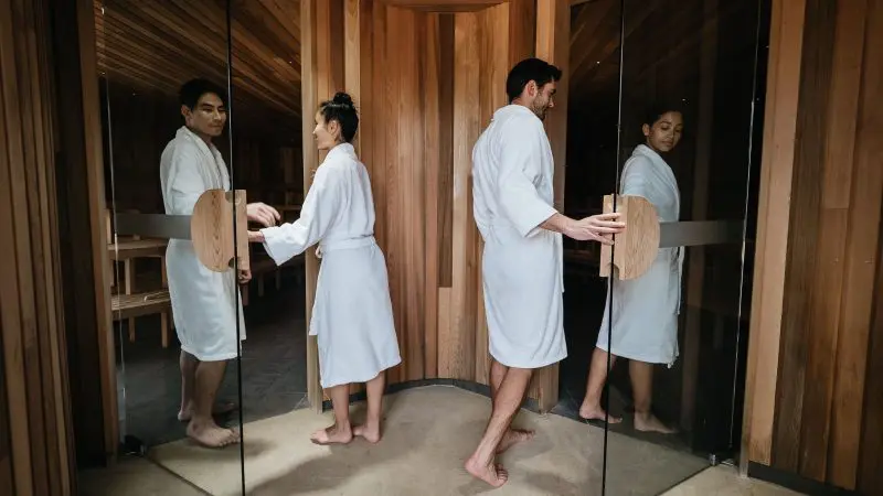 Two guests in white robes enter a wooden sauna at Peninsula Hot Springs, holding 1 Day Spa Entry shuttle bus passes for ultimate relaxation.