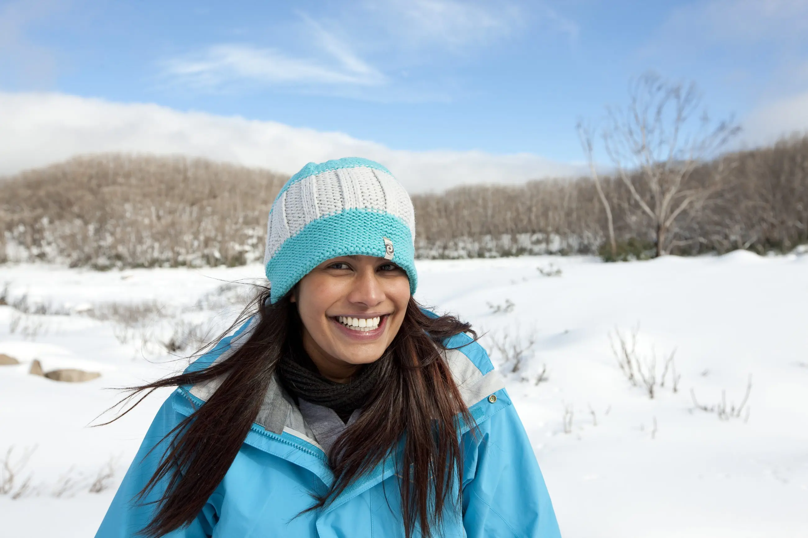 Happy woman in blue jacket enjoying a 1 Day Lake Mountain Snow Waterfalls Tour from Melbourne, with scenic snowy trees behind her.