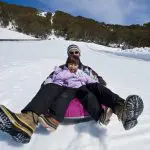 Smiling adult and child in warm winter gear sledging down a snowy hill on a Lake Mountain Snow Tour, near Melbourne, Australia.