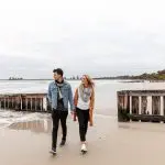 A couple walks along a scenic sandy beach with wooden barriers, capturing the romance of a Great Ocean Road Reverse Eco Tour adventure.