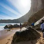 Group of four tourists on a scenic sandy beach, backed by dramatic cliffs, during a top-rated 1 Day Great Ocean Road Rainforest Tour.