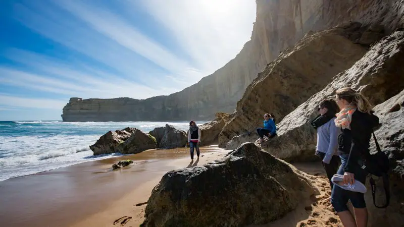 Four tourists relax on sunlit rocks at a sandy beach during a popular 1 Day Great Ocean Road Reverse Eco Tour, enjoying scenic views.