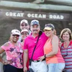 Group of six happy adults pose by the iconic Great Ocean Road sign on a 1 Day Reverse Eco Tour, enjoying scenic Australian adventure.