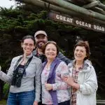 Group of four happy adults stand beneath the iconic Great Ocean Road sign on a 1 Day Great Ocean Road Rainforest Tour in Australia.