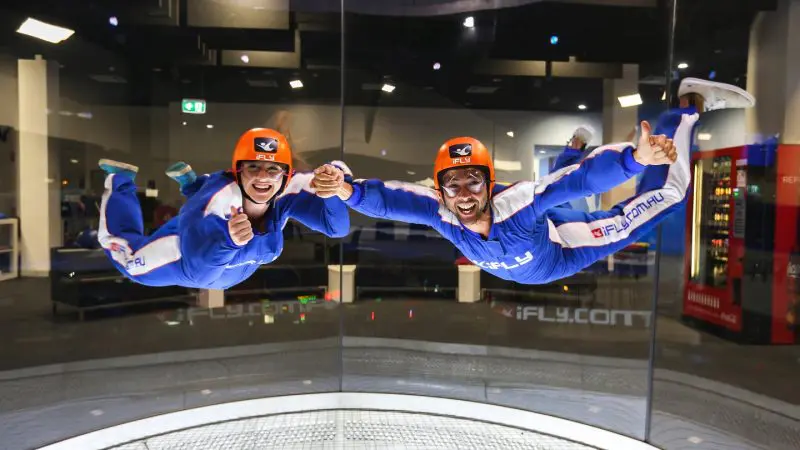 Two people in blue jumpsuits and orange helmets, smiling as they experience an indoor skydiving session at iFLY Family and Friends.