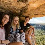 Three happy travellers on a 1 Day Blue Mountains Wildlife Park tour, smiling atop a rocky ledge with panoramic views of lush green valley.