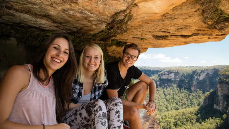Three happy travellers on a 1 Day Blue Mountains Wildlife Park tour, smiling atop a rocky ledge with panoramic views of lush green valley.