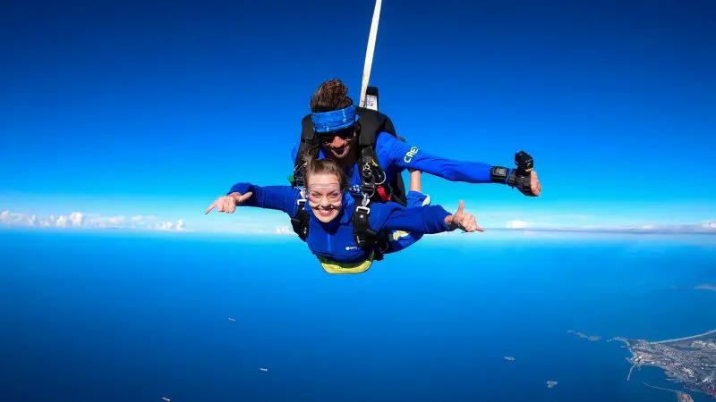 Two thrill-seekers experience a Newcastle tandem skydive at 15,000ft, grinning with arms spread wide above the sparkling blue sea.