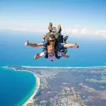 Tandem skydivers above Byron Bay coast, smiling and giving thumbs up during 15,000ft skydive with Brisbane transfer included.